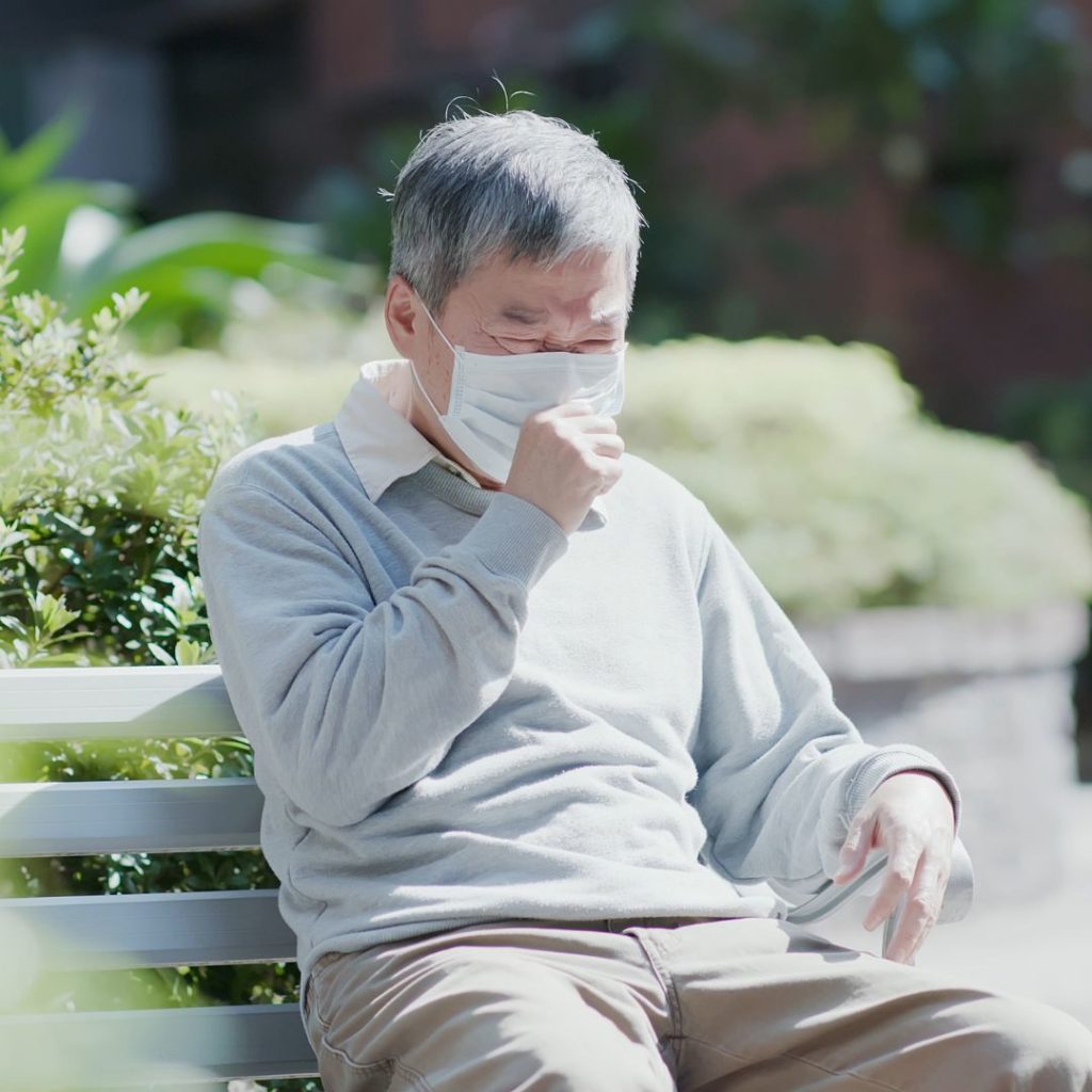 Elderly man sitting on a bench wearing a mask holding his hand up to his mask because he is coughing